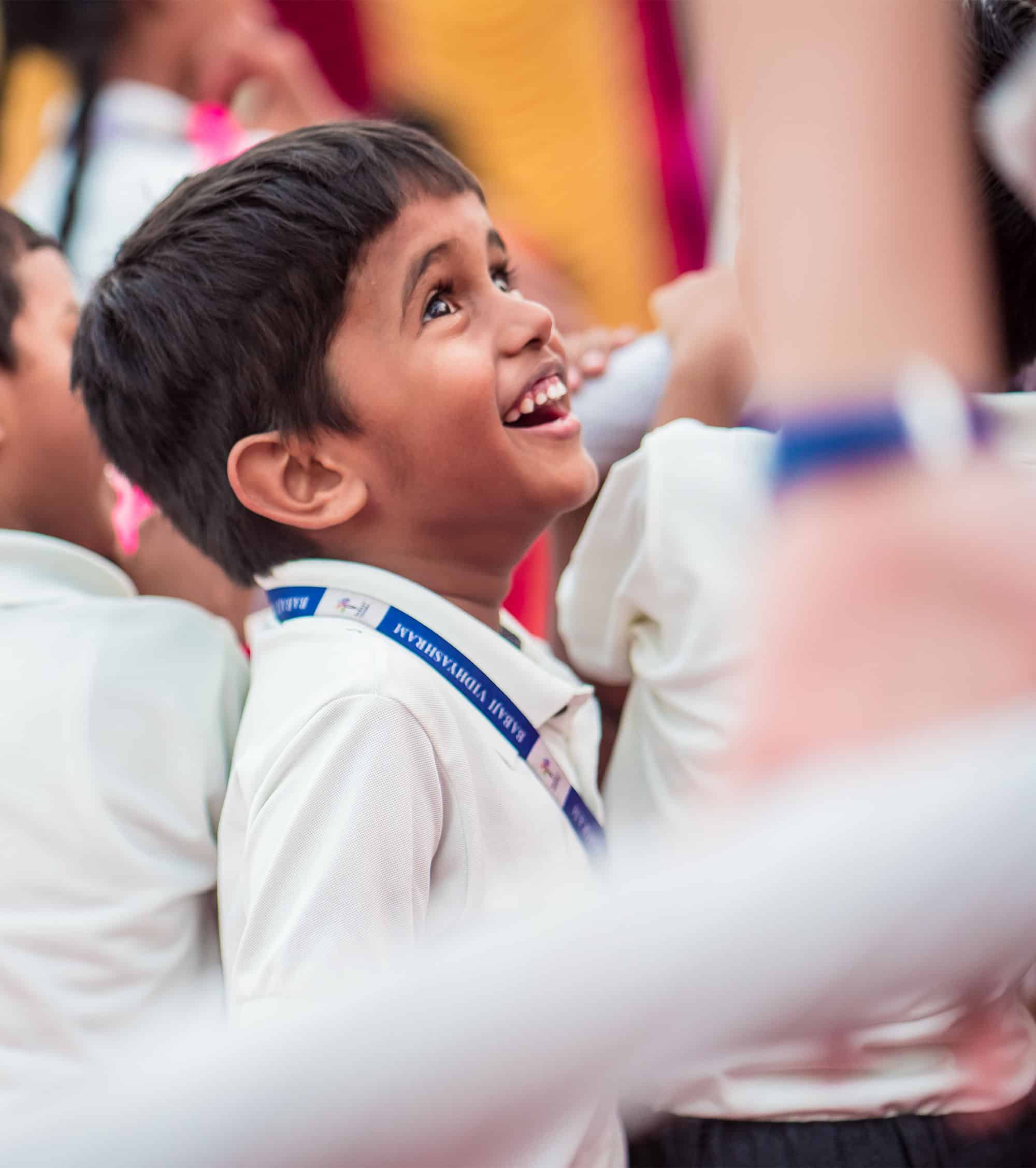 A small school boy with a school uniform and identify card on his neck is having a broad smile on his face.